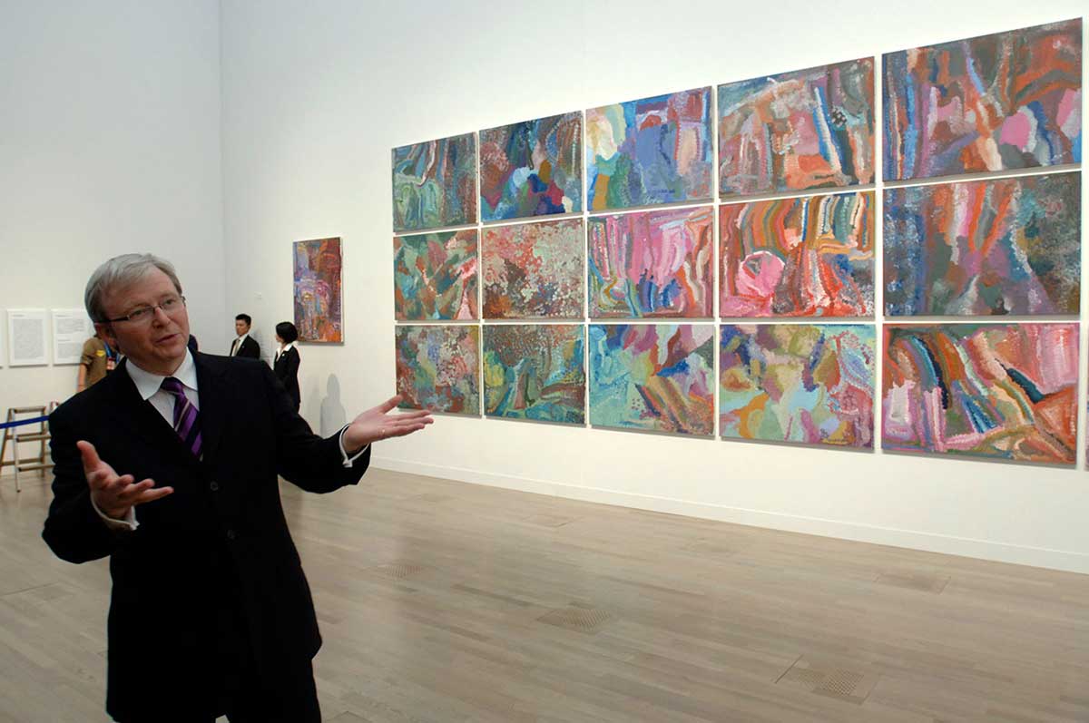 A man standing in an exhibition space with a large painting mounted on the wall in the background. - click to view larger image