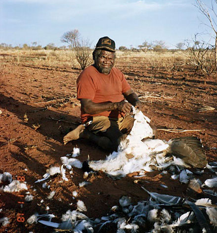 Aboriginal Australian man in an orange T-shirt and cap plucking a bird. - click to view larger image