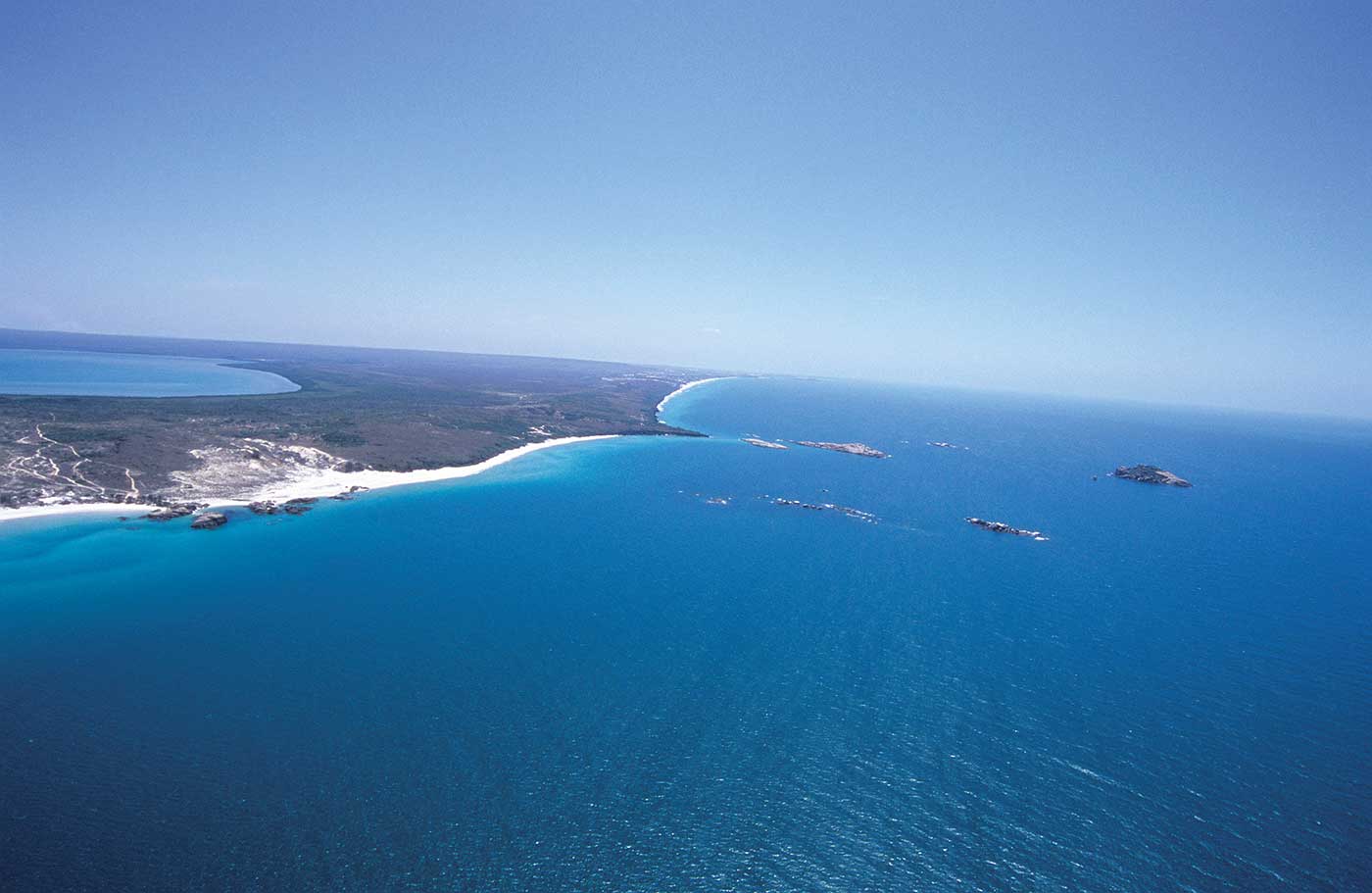 A colour photograph of part of a peninsula. The photograph has been taken from an aeroplane. The peninsula is to the left of the photograph; it has a white sandy beach on the coastline closest to the camera. In the foreground, stretching back to the horizon, is the ocean, which is a deep blue colour. The sky is in the upper part of the photograph; it is a more pale blue in comparison to the ocean.