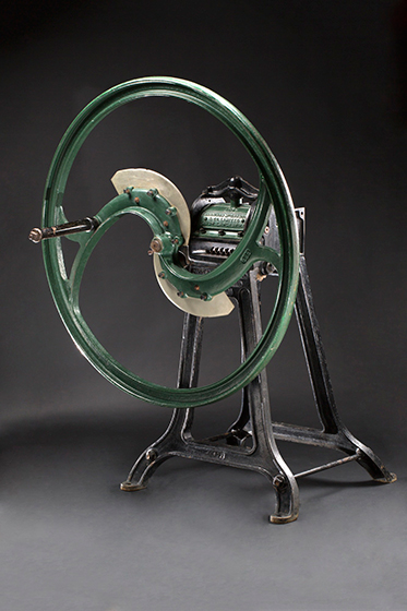 A piece of metal farm machinery with a large circular wheel, with a handle attached to the front, atop a four-footed stand. - click to view larger image