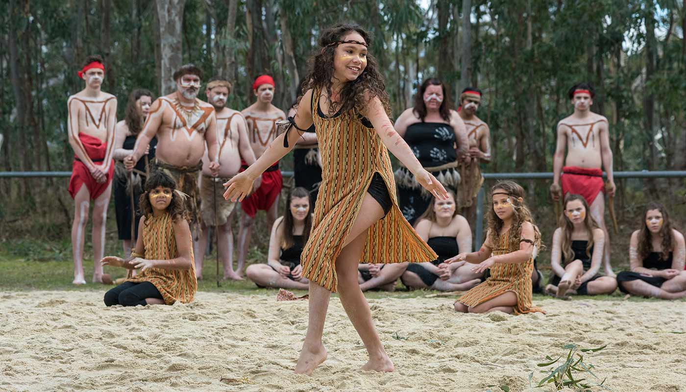 Australian Indigenous dancers perform on sandy ground with a forest background behind them.