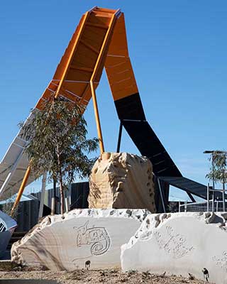 View of a large sculptural architectural feature rising above landscaped grounds of native plants. In the foreground there are large stones with etchings of Australian wildlife. - click to view larger image