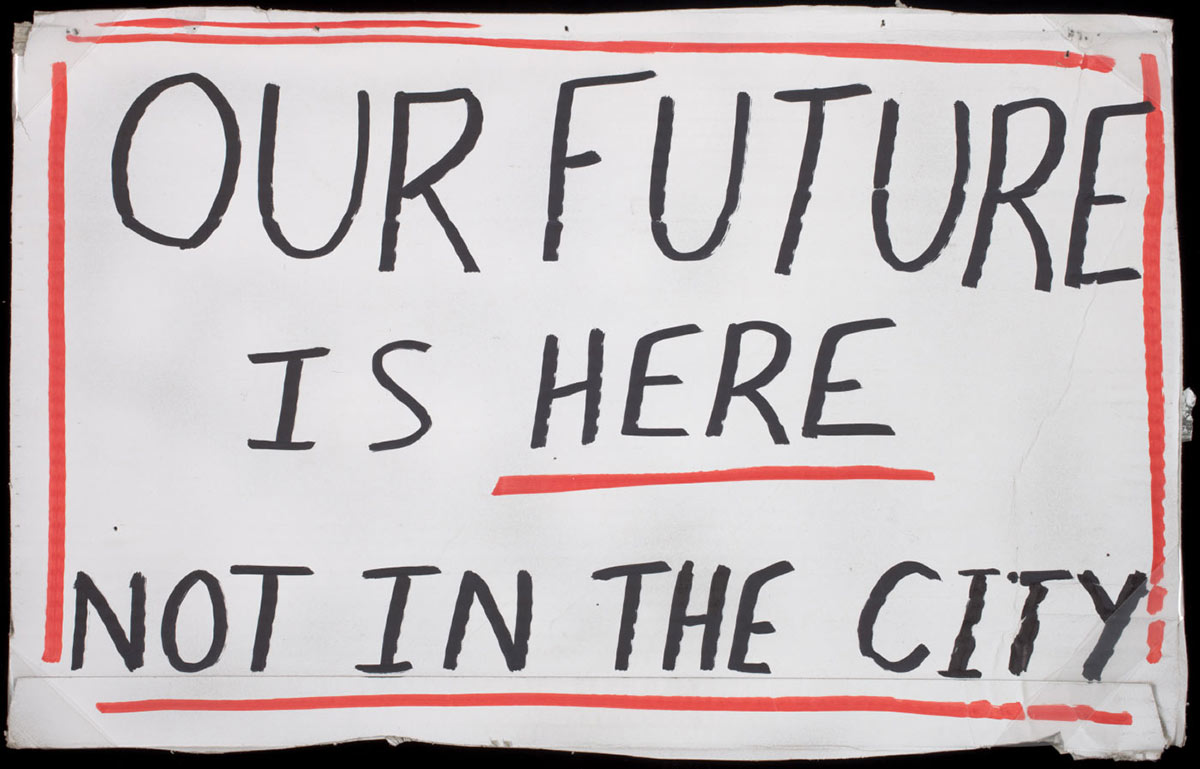A placard made from a cut out of a cardboard box. There is black handwritten text against a white background on the front surface that reads 'OUR FUTURE / IS HERE / NOT IN THE CITY'. The text is surrounded by a drawn on red border and the word 'HERE' is also underlined with a red line. There is a yellow sticker on the back of the placard with the printed words 'BOURKE FURNITURE ONE / ...'.