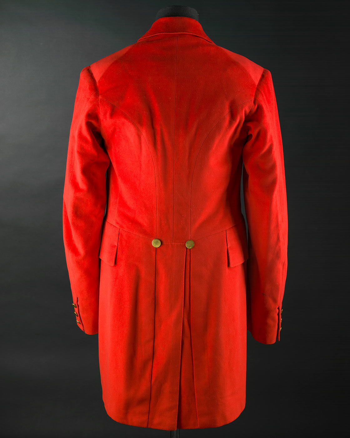 A red wool cut away jacket with tails otherwise known as hunting pinks. It has 3 brass shank buttons at front opening and 3 buttons on each cuff. It is fully lined in red cotton. The sleeves are lined in white cotton with blue stripe. The jacket has machine quilted underarm lining and a back vent with 2 brass buttons at top. It has four pockets on front with flaps. - click to view larger image