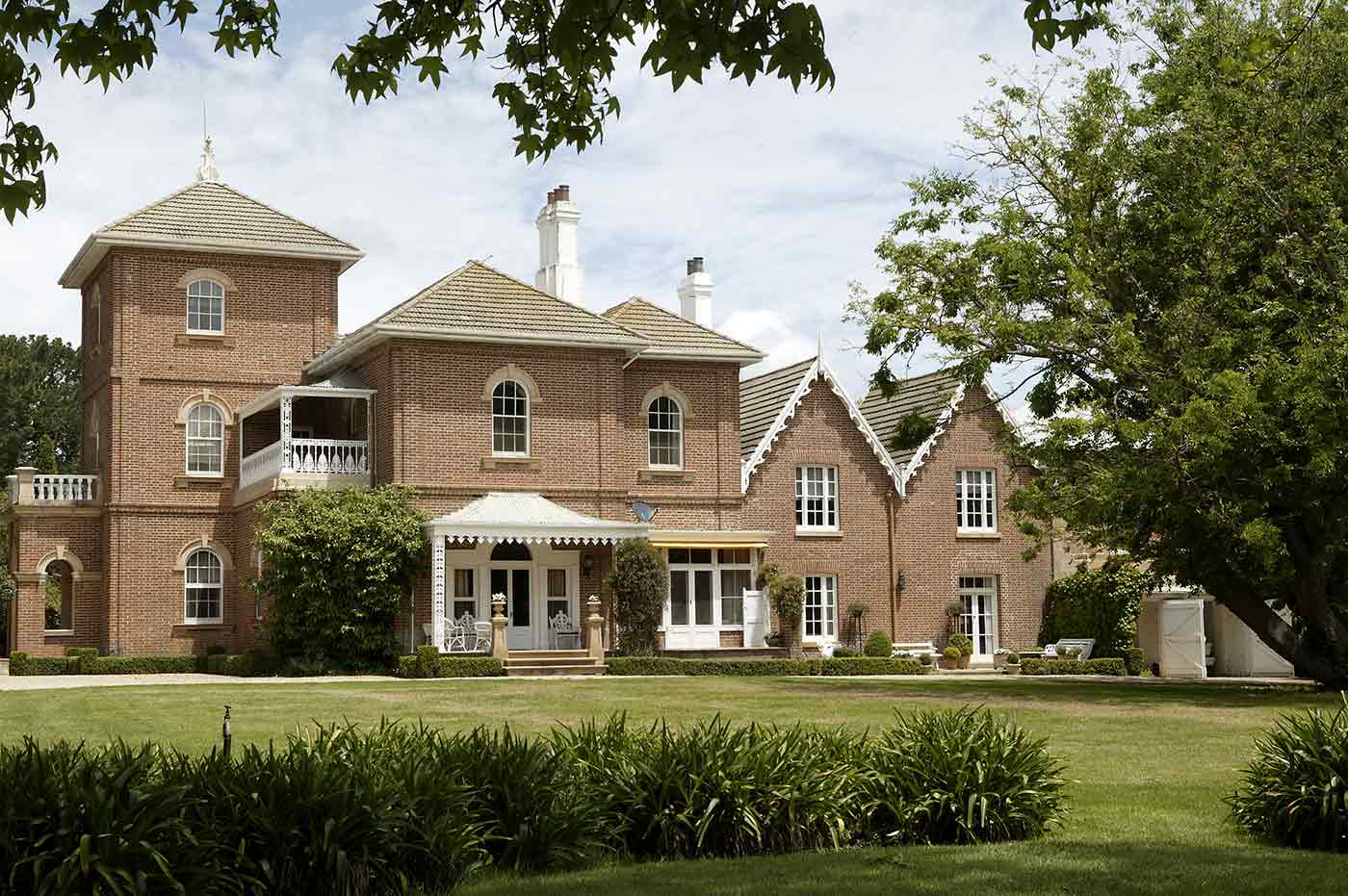 Colour photograph showing part of a large, two-storey brick house with tower, white chimneys and extensive gardens.