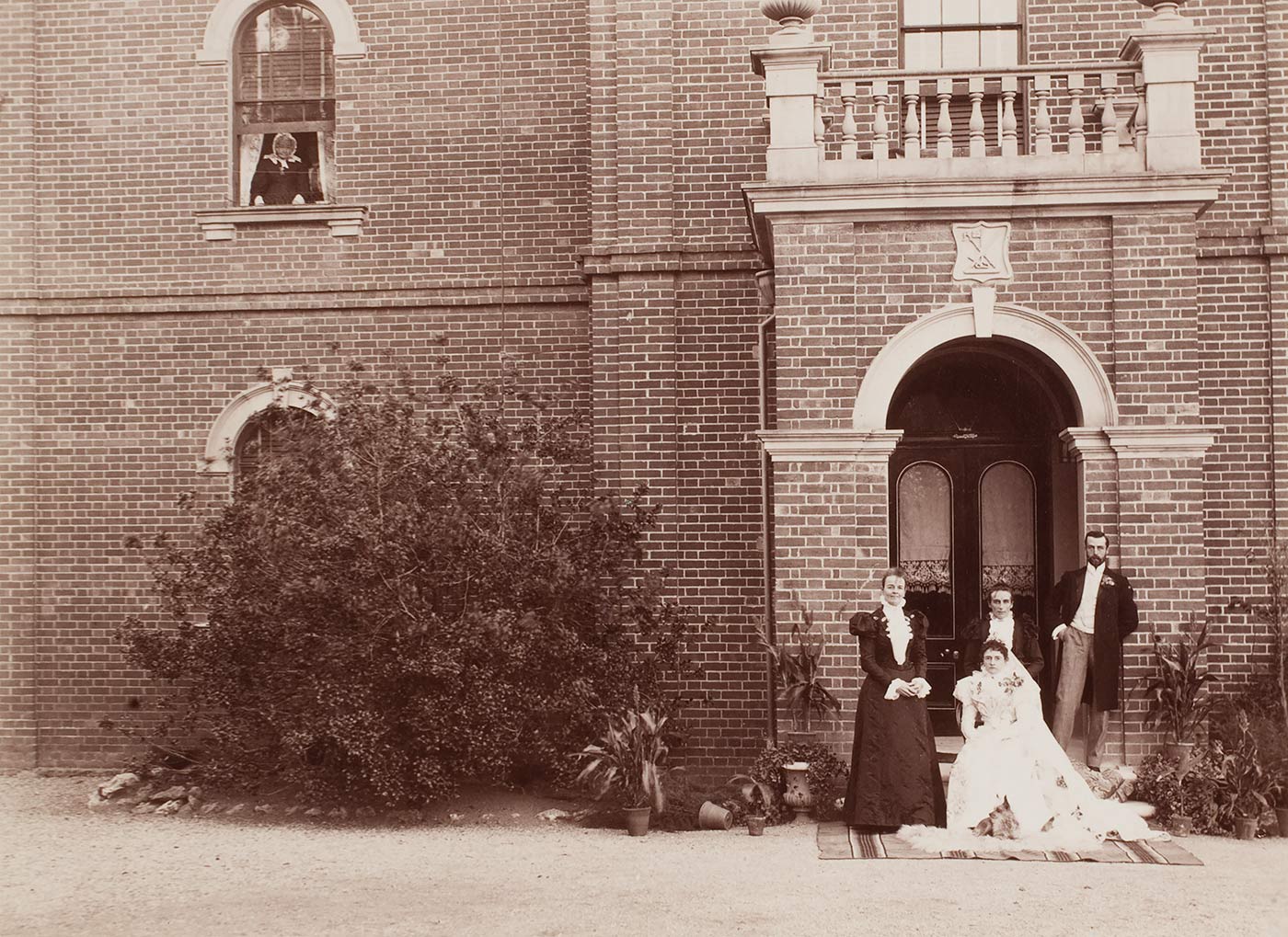 A black and white photograph that shows a wedding party standing below an arched entrance way in front of a section of a brick building. There is a bush to one side and a figure looking out of a window in the upper left section of the photograph. - click to view larger image
