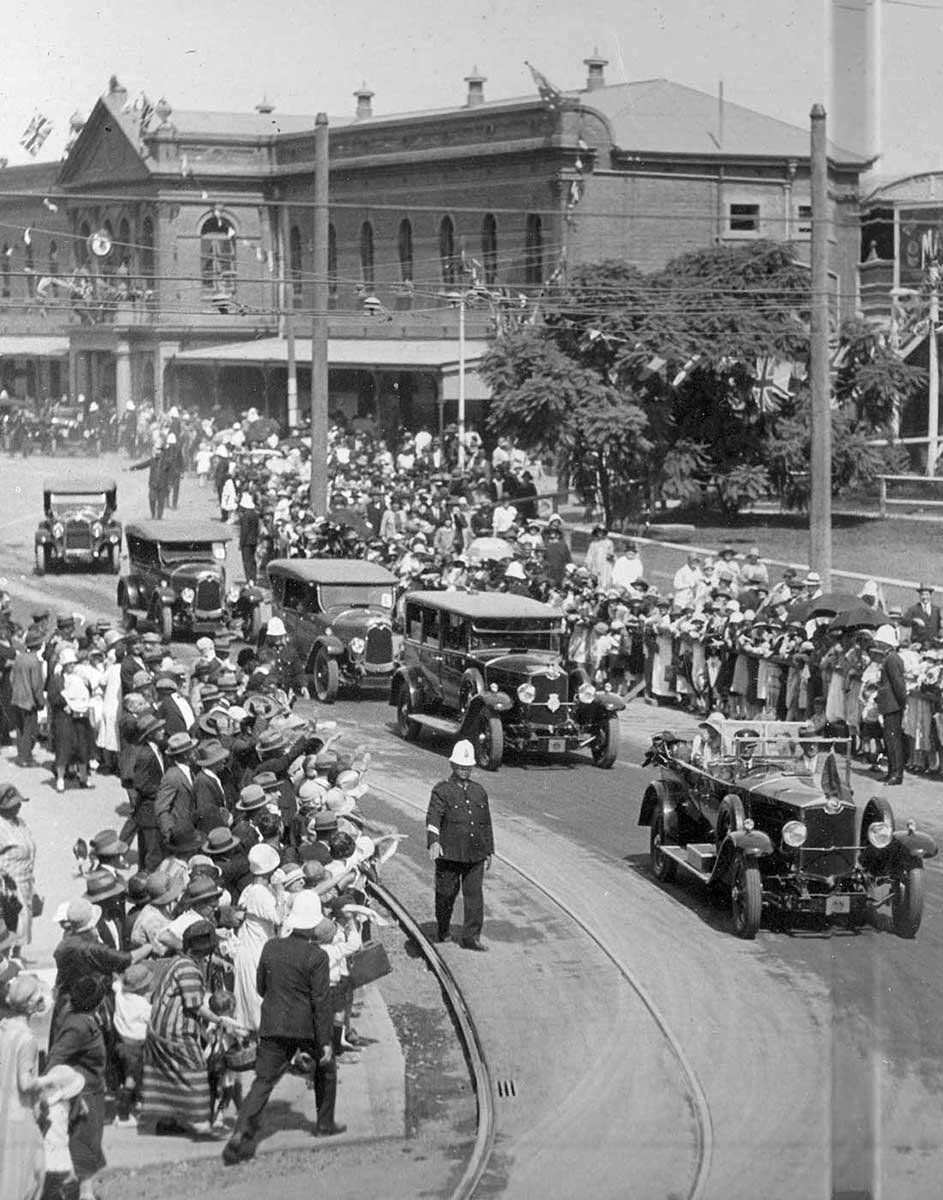 A black and white image showing five cars in a procession travelling past the South Brisbane Railway Station. Crowds line the streets with uniformed police officers patrolling. - click to view larger image