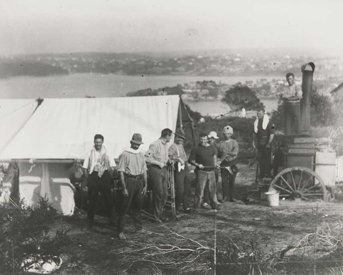 A black and white photograph of nine men standing outside a tent and next to a portable boiler. Some of the men have towels aroudn their necks and one is holding a long handled saucepan. Suburbs are visible on the headlands in the distance. - click to view larger image
