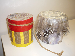 Two decorated drums. One is decorated with cardboard and red and yellow tape, the other is decorated with string and feathers.