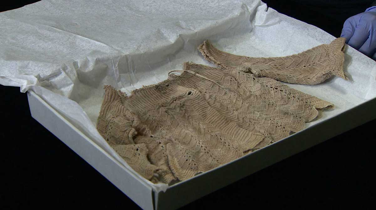 A stained, knitted baby's jacket in a white box. The jacket has heavy soiling around the neckline and the left sleeve has been ripped off. - click to view larger image