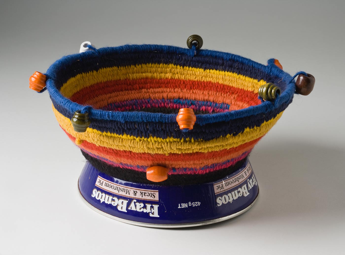 A circular coiled multicoloured yarn and plant fibre basket with a metal base and bead decoration. The base is made of a blue metal 'Fray Bento / Steak and Mushroom Pie' tin with sides, that is placed upside down. The yarn section is attached by black yarn threaded through holes punched in the metal. The closely coiled yarn is in stripes starting with blue at the top edge then yellow, red, orange, pink and black. The top edge and sides have a decoration of evenly spaced carved cylindrical wooden beads in orange, brown, green and white. - click to view larger image