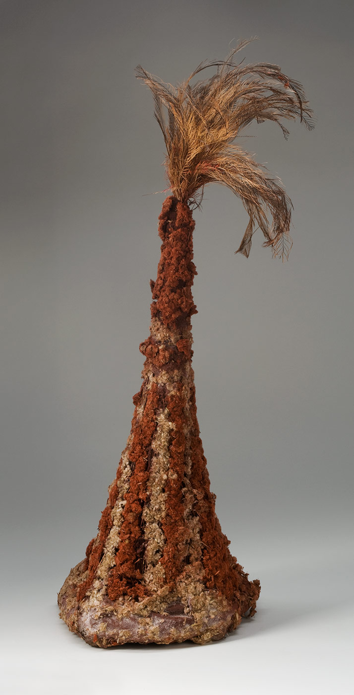 A tall conical headdress made of plant fibres attached to a firm circular ring at the base and covered with pigmented fluffy plant fibre with feathers at the top. The ring at the base is pigmented red and the plant fibre stalks are attached around it. The fibres are bunched together towards the top and wrapped with maroon coloured string. The plant stalks are covered with fluffy plant fibre which is decorated with red pigment in vertical stripes on the bottom half and red at the top. At the top of the headdress is a bunch of fluffy and spiky brown, tan and grey feathers. - click to view larger image