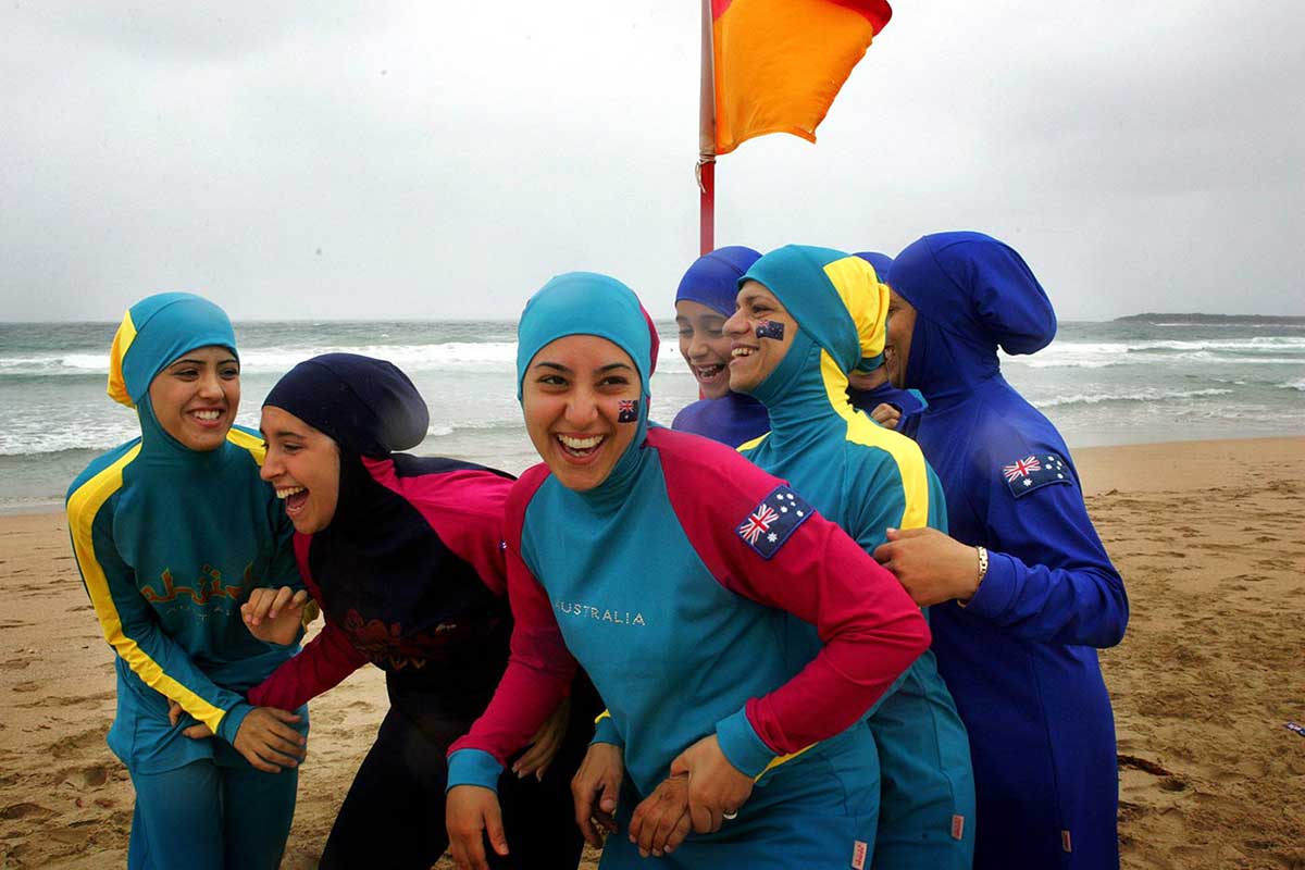 A group of young women of middle-eastern appearance laughing and embracing each other. They are wearing colourful beach wear that has been designed to include the burqa.