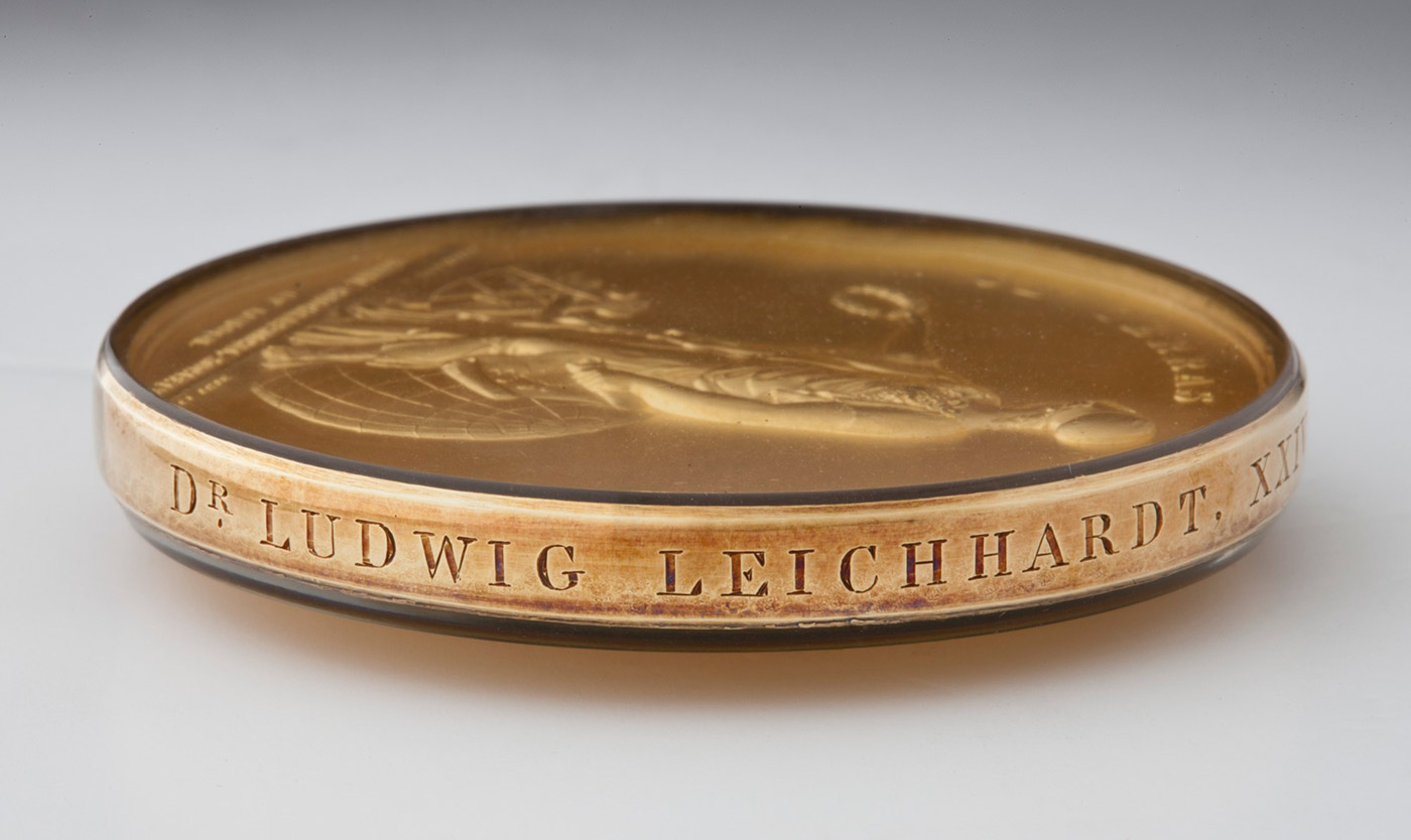Compile of three images showing a circular, gold-coloured medal at left, a close-up image of an outstretched arm holding a laurel wreath, top right, and the letter 'LU' on a scratched surface, bottom right. - click to view larger image