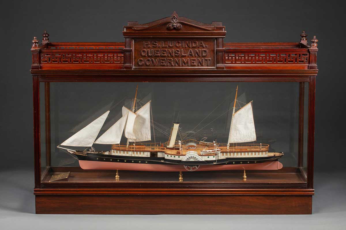 A model of a ship in a wood and glass case, the ship has five raised sails, water wheels on either side, painted in black and brown with details in blue and red, held on four gold coloured stands, and a gold plaque at one end. Th case is in good condition and has an engraved panel with the text: 