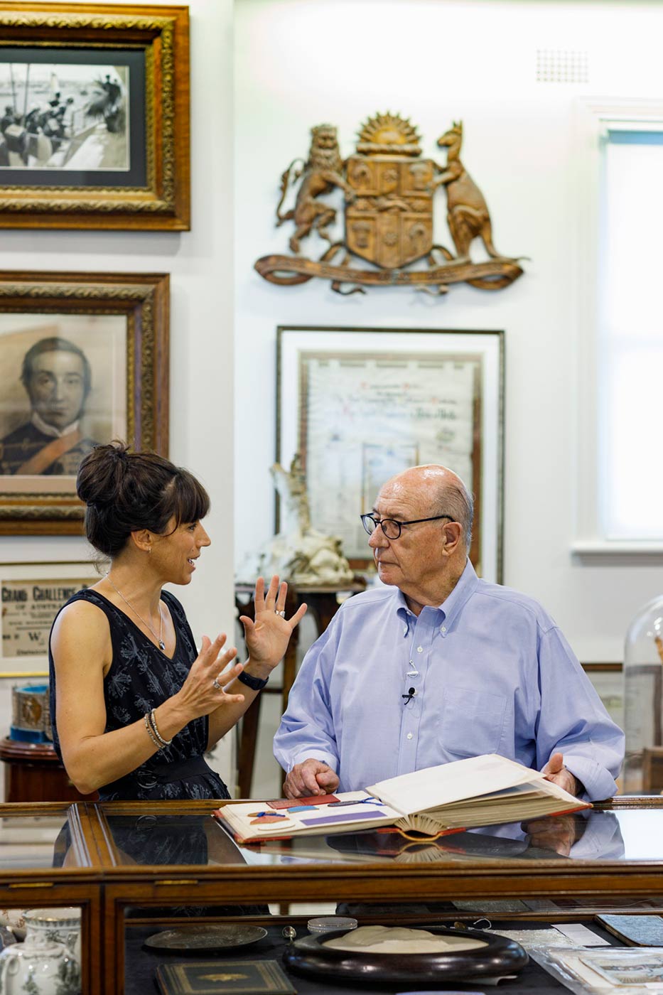 Colour photograph of a woman and man in discussion in a room featuring various antique collectible objects. - click to view larger image