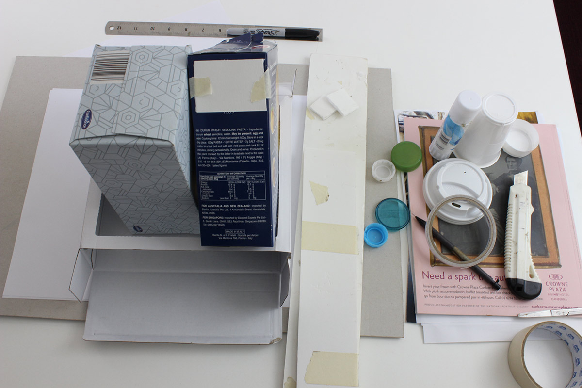 Various materials for a craft project including cardboard boxes, plastic lids and cups, glue, tape, ruler, pen, etc.