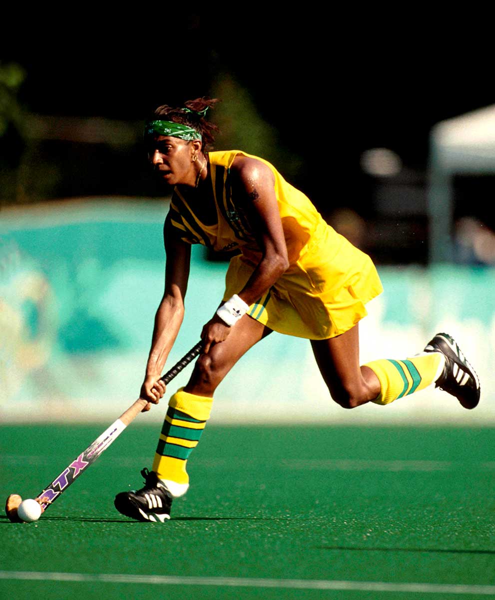 A woman in a green and yellow uniform running with a hockey stick across a field. - click to view larger image