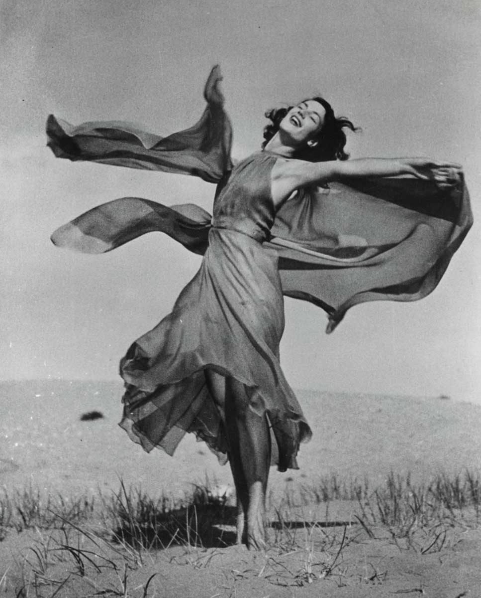 Black-and-white portrait photograph, showing a smiling woman dancing outdoors in a sandy landscape. Her arms are extended, and she appears to be spinning, her dress flowing and billowing around her.  - click to view larger image