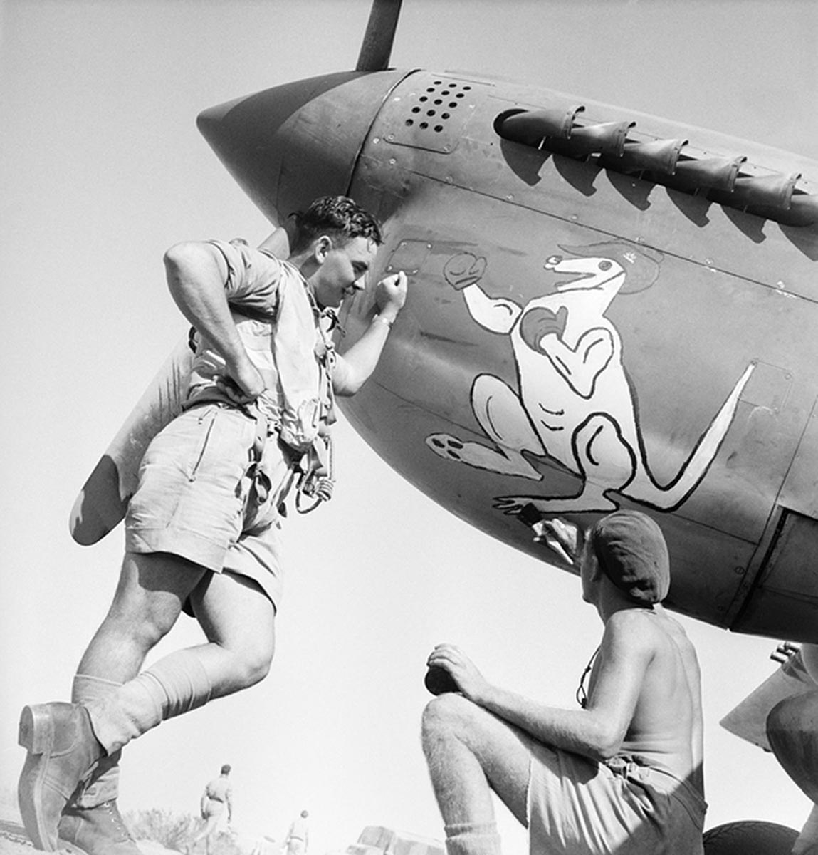 A black and white photograph taken at Agnone. Flying Officer D Davidson (left) of Sydney, NSW, looks on whilst his fitter, 16528 Leading Aircraftman Jack Ronald Clifford Featherby (right) of Sydney, NSW, puts the finishing touches to some nose art, a boxing kangaroo, which some (possibly a flight), of the 'Desert Harassers' of No 450 (Kittyhawk) Squadron, RAAF, have adopted as an insignia during operations over Sicily and Italy. - click to view larger image