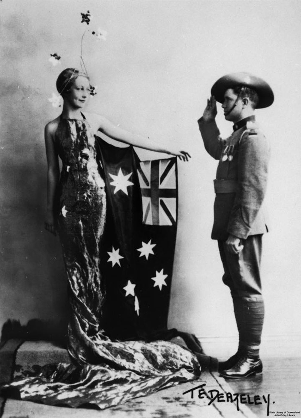A woman dressed in a sleeveless fitted evening gown, which trails to the floor, wears a headdress of stars. An Australian flag is attached to her left arm, which is extended in the direction of a young man. He wears a soldier's uniform and has his right arm raised in a salute. - click to view larger image