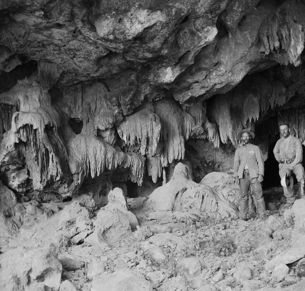 Two non-Aboriginal men standing at the entrance to Wangalinnya Caves, Napier Range, Western Australia 1916. The cave entrance is made up of fantastic rock formations rising from the ground and hanging from the entrance roof. The formations hang in frond-like formations or appear to grow from the ground. The two men stand to the far right of the image.
