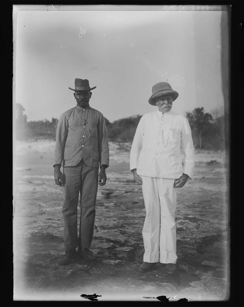 A positive print of two men. One man is Aboriginal and the other non-Aboriginal. They stand next to each other on open ground; the Aboriginal man is to the left in the image. He wears shoes, trousers, a shirt and a hat. The other man wears shoes, trousers, a shirt and a pith helmet. Both stand in stiff, formal poses with their hands by their sides. The image is in black and white. - click to view larger image