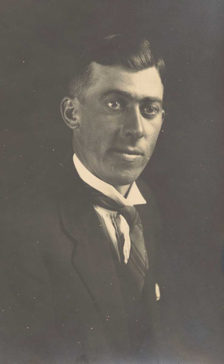 Portrait photo of Frank Feast, about 1920s. He sits facing toward the right side of the photograph, with his head turned slightly toward the camera. He wears a shirt with upturned collar, tie and jacket. He is visible from about mid-torso height up. His head is slightly inclined toward his left shoulder. Mr Feast has his hair slicked back in the popular style of the era. It is short at the sides. The photograph appears to have been taken in a studio, as the dark, featureless background and strong light source from beyond the right side of the image suggest this setting. - click to view larger image