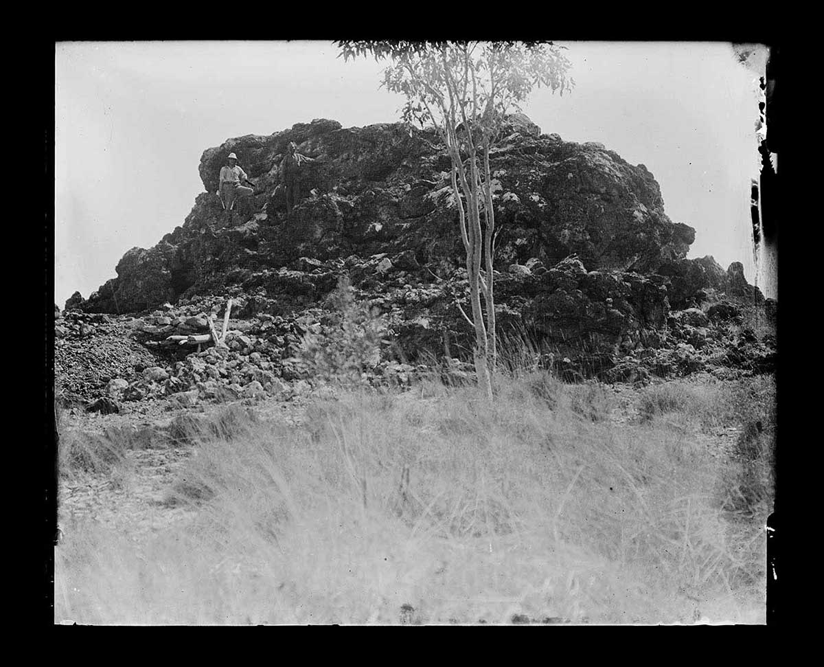 Two men standing on a rocky outcrop, Narlarla, Napier Range, Western Australia 1916. The outcrop is about six metres high, with a rough surface and signs of significant erosion. The two men stand toward the left of the outcrop. They are hard to see in amongst the shadows of the outcrop's surface. A single young tree is in front of the outcrop, in the middle distance. - click to view larger image
