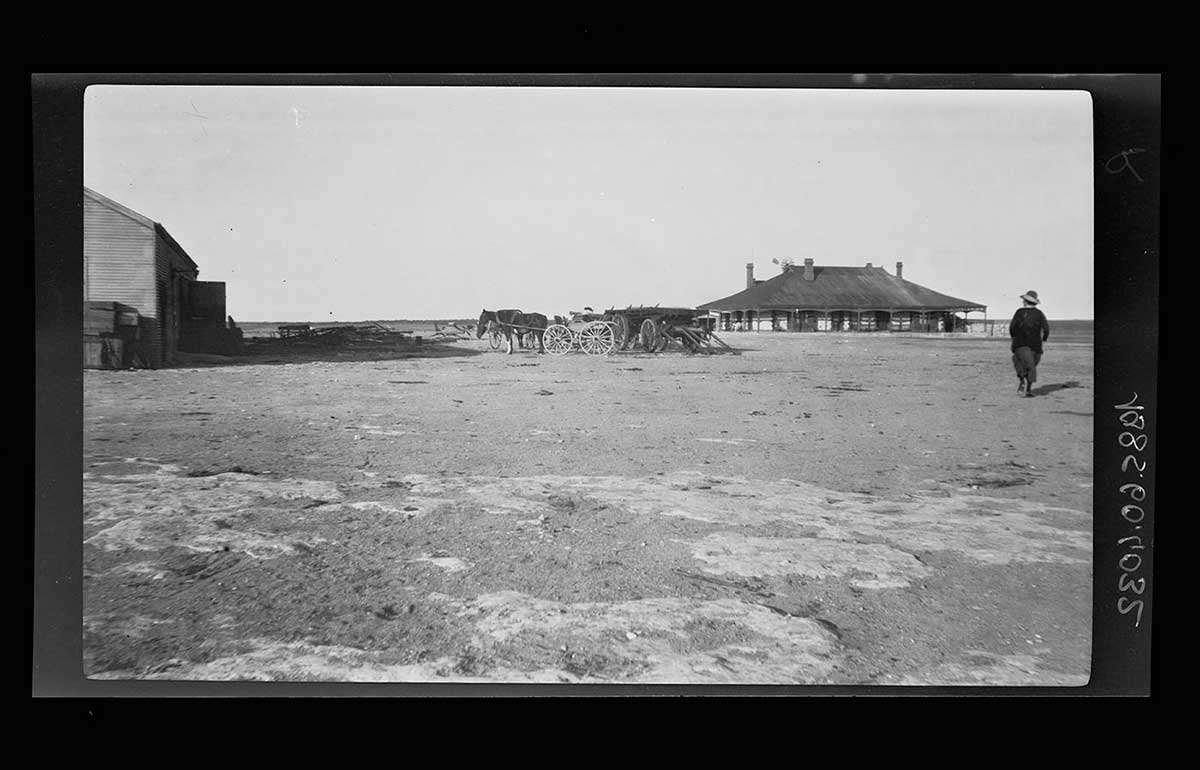 Yalata station homestead, South Australia 1920. The homestead is in the right far distance of the image. A woman in the right middle ground walks toward it with her back to the camera. Two drays with horses are in the middle of the image. The corner of a large barn style building is at the left side of the image. The foreground shows bare earth with what appear to be exposed sections of flat rock. - click to view larger image