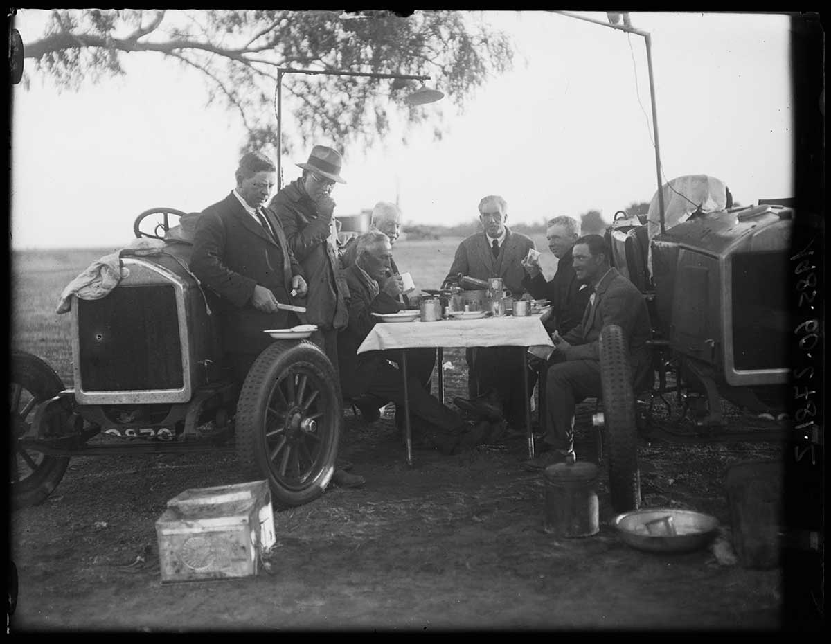 The vice-regal party at breakfast, central Australia 1923. Seven non-Aboriginal men sit around a small table between two cars. On the table are plates, mugs and food. A tablecloth covers the table. Two of the men at left lean against the car on that side while two next to them sit on the car. One man appears to be sitting on a chair at the back of the table. The two men on the right sit on the car on that side. All wear suits; one wears a Trilby-style hat. - click to view larger image