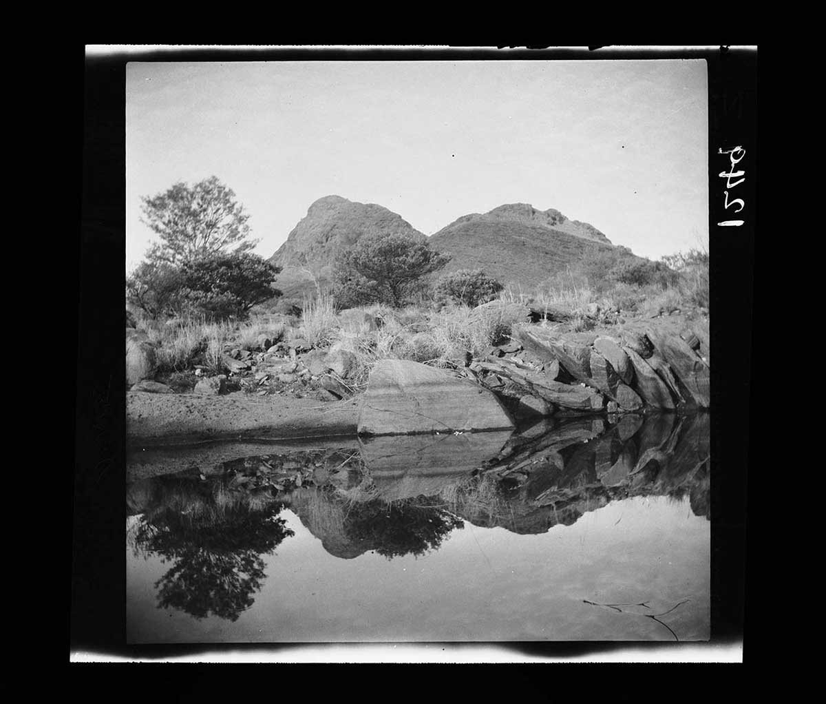 Waterhole at foot of Ruined Rampart, Petermann Ranges, Northern Territory 1926. The waterhole is in the image foreground. Its surface is still and mirror-like, reflecting the landscape and the sky. In the middle ground at the water's edge are numerous large rocks, some stacked next to each other like books. A large smooth rock in the image centre disappears into the water. Beyond the rocks is scrub with scattered trees and bushes. In the distance is a mountain range, with two ridges in front and a third ridge behind them. - click to view larger image