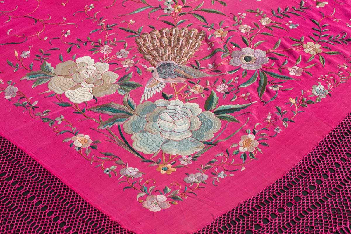 Photograph of corner details of a pink silk shawl. The edges of the shawl have a hand-woven honeycomb pattern and long fringed edge. The corner of the shawl has large embroidered floral motifs, with three larger flowers with gold, blue and purple silk thread, and an embroidered peacock-like bird. These are surrounded by smaller, more intricate floral details and vine patterns. - click to view larger image