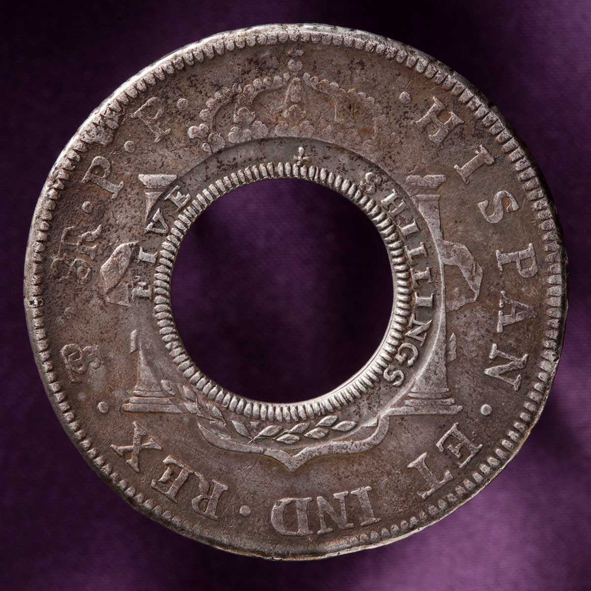 Studio photo of silver coin with missing centre. The original Spanish imprinting in Latin is visible around the outside rim, while the words five shillings have been imprinted around the inside rim. - click to view larger image