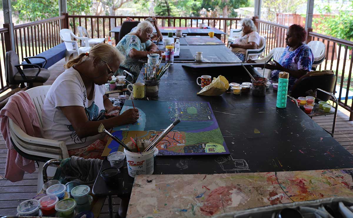 Five people are painting together around long tables laden with canvases, and other other art materials. - click to view larger image