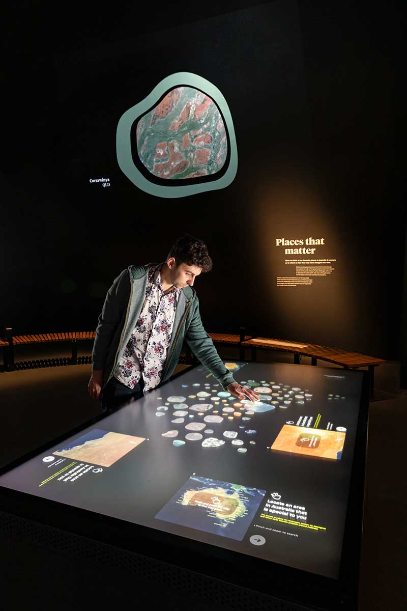 A man stands beside a backlit screen in a museum gallery. He is touching a point on a map. An image of the earth from above is projected on the wall behind. - click to view larger image