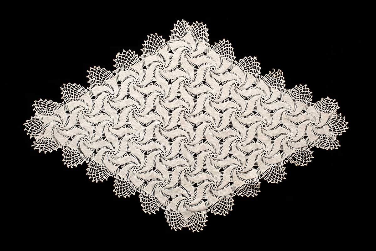 Diamond-shaped crocheted table setting in white thread on a black background. Table setting has a repeated spiral pattern and scalloped edges. - click to view larger image