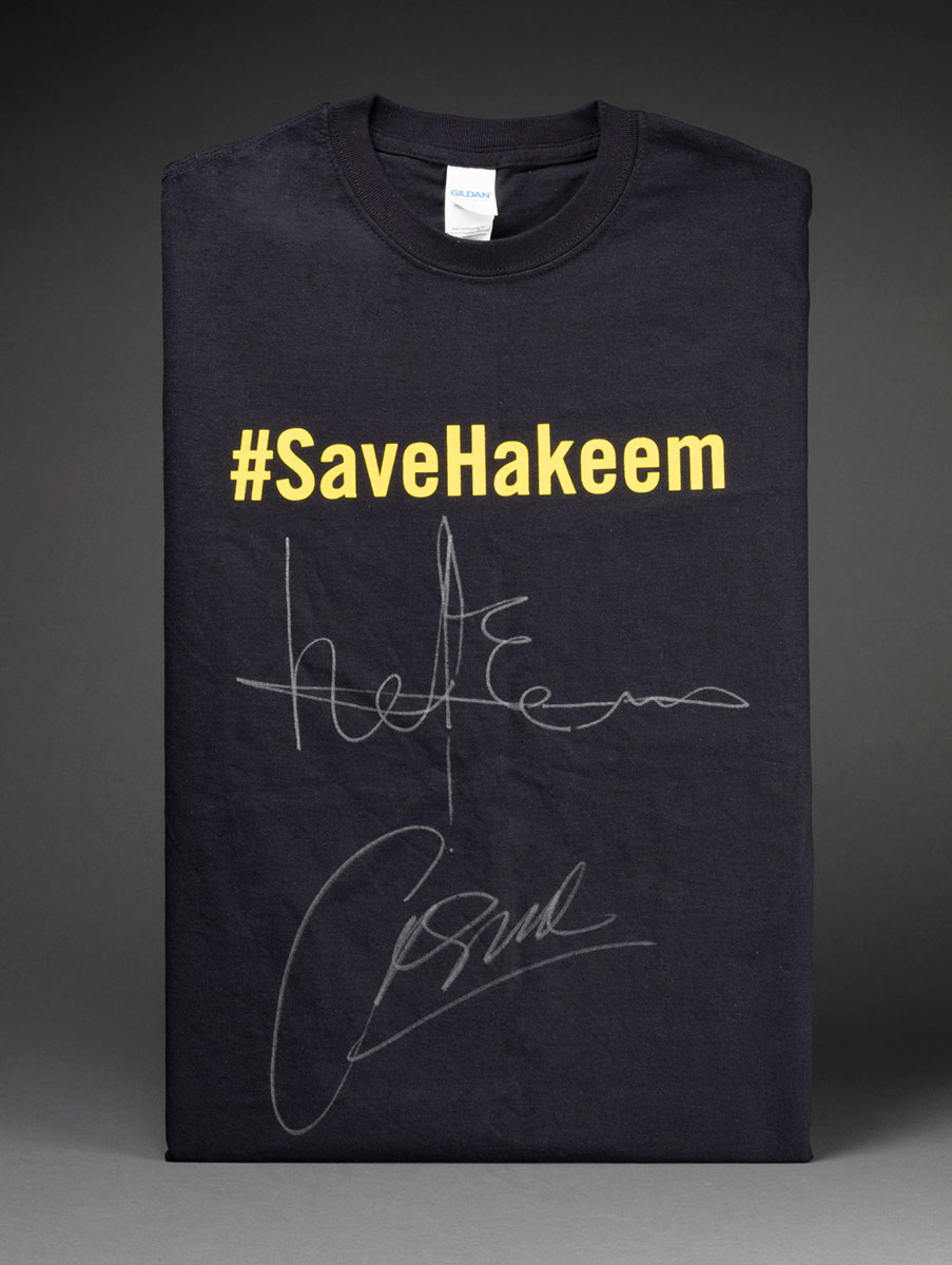 A black, cotton t-shirt with '#SaveHakeem' painted in yellow across the chest. Below this are two signatures written in white.
