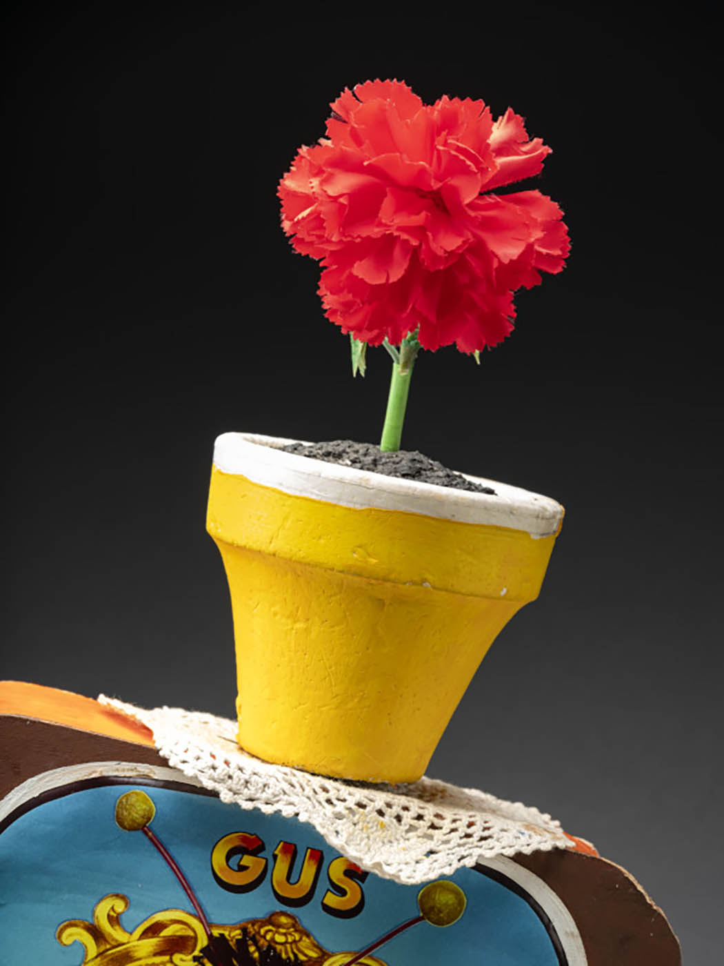 A puppet of a snail named Gus. Gus has an orange body, a tuft of black hair, orange and yellow antennas, brown eyes and a pink felt tongue. Attached to its back is a television box with two knobs, a screen featuring an image of Gus’s head surrounded by a gold frame, and printed text that reads ‘GUS/TV’. Attached to the top of the television is a yellow flowerpot with a red flower, sitting atop a doily. 