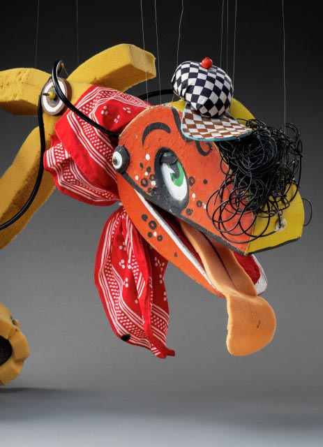A puppet named Bill Steamshovel, in the form of an excavator. Bill Steamshovel consists of a red, yellow and green wooden control room connected to which are foam tread tyres and a foam neck. At the end of the neck is a red and yellow excavator head with a checkered hat and hair made from strands of black rubber. Tied around the neck is a red and white handkerchief. 