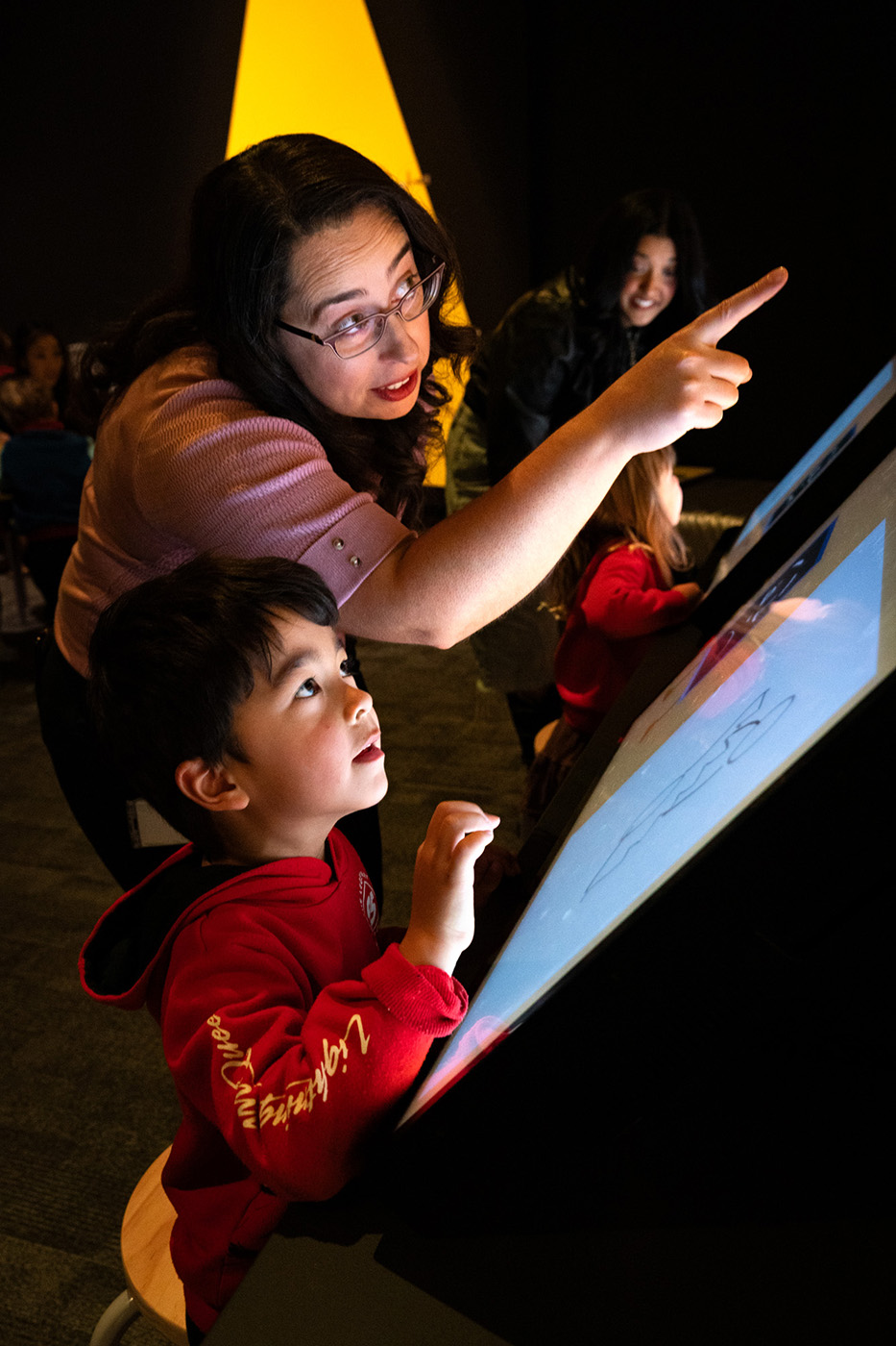 A woman stands, pointing at an unseen object, beside a young child, who looks up. 