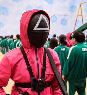 A man in a hooded pink tracksuit, with a black face mask with a white triangle symbol on the forehead, stands amid a field of people, some wearing green tracksuits, unmasked, and others wearing black face masks and pink tracksuits.