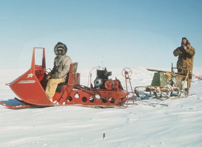 A bright orange-red, open-topped powered snow vehicle in an Antarctic landscape. The driver, seated at the front, wears a hooded jacket and dark goggles, with only his nose visible. Another person stands to the rear. The vehicle tows a sled with scientific equipment on top.