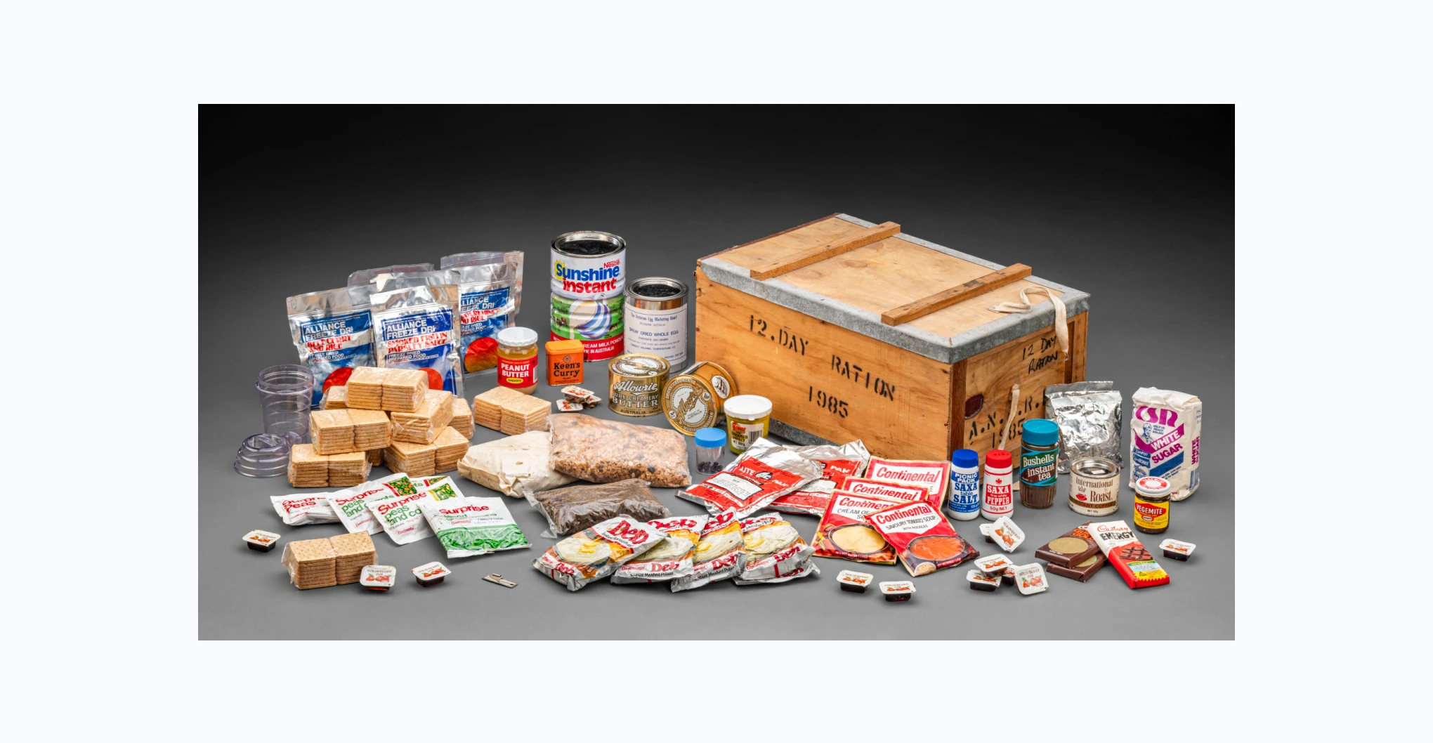 A wooden crate with a hinged lid containing various rations. The edges of the lid are reinforced with metal and the lid can be secured closed via two lengths of cotton tape. Stencilled in black on the crate is '12. DAY RATION / 1985' and 'A.N.A.R.E. / 1985'. There is also handwriting in black that reads '12 DAY / RATION'. The rations include a salt shaker, a pepper shaker, four satchets of Continental freeze dried soup, 10 packets of Swallow's biscuits, six packets of Alliance freeze dried meals, two packets of Alliance freeze dried rice, two packets of dried onions, five packets of Continental dried vegetables, a jar of Vegemite, a container of Keen's curry powder, a tin of instant coffee, a tin of Nestlé milk powder, a tin of dried egg powder, two tins of Allowrie butter, three blocks of Cadbury chocolate, a bag of dried fruit, a bag of cereal, a bag of muesli, a bottle of Bushells tea powder, a jar of smooth peanut butter, a jar of 12 black pills, a container of creamed honey, a bag of CSR sugar, 15 packets of Kraft strawberry jam, a plastic shaker bottle, a metal can opener, and five packs of Deb instant mashed potato.