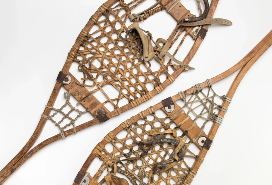 Detail view showing two snowshoes, side by side from above, with pale wooden frames, in elongated teardrop shape, with crisscrossed leather webbing, and metal reinforcements at either end.