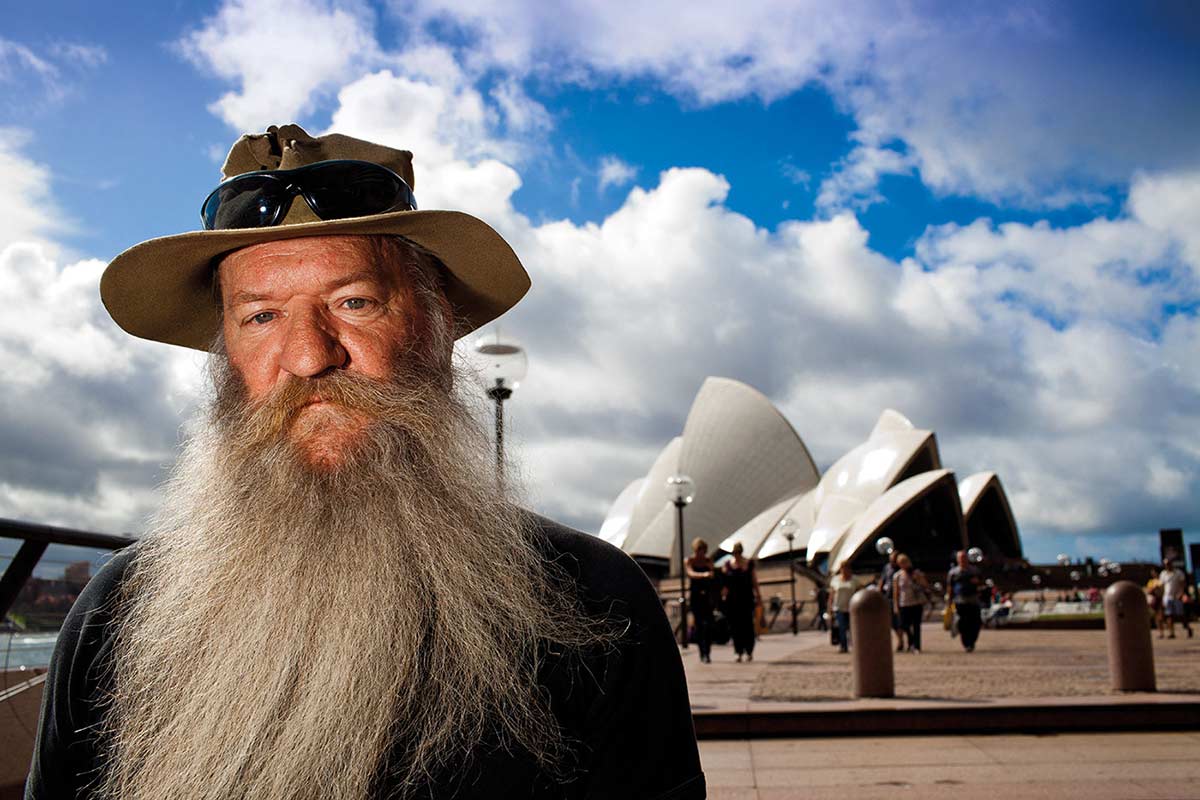 Portrait of a man with a long grey beard, and wearing a khaki hat with sunglasses on top, standing to the left, with a distant view of the Sydney Opera House, people walking on a concourse, and a blue sky with white clouds.