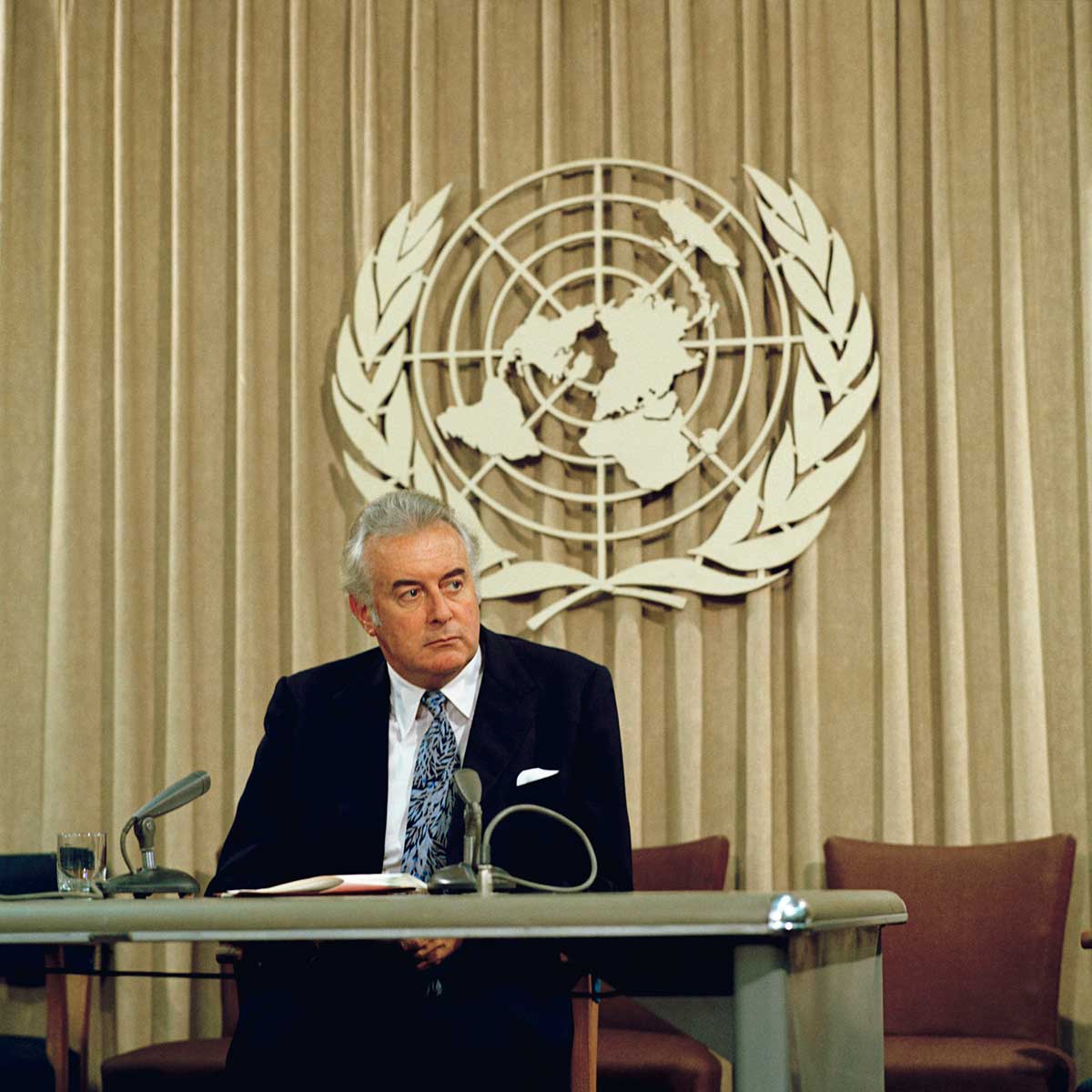 Colour photo of middle-aged man in suit sitting behind desk with UN symbol in background.