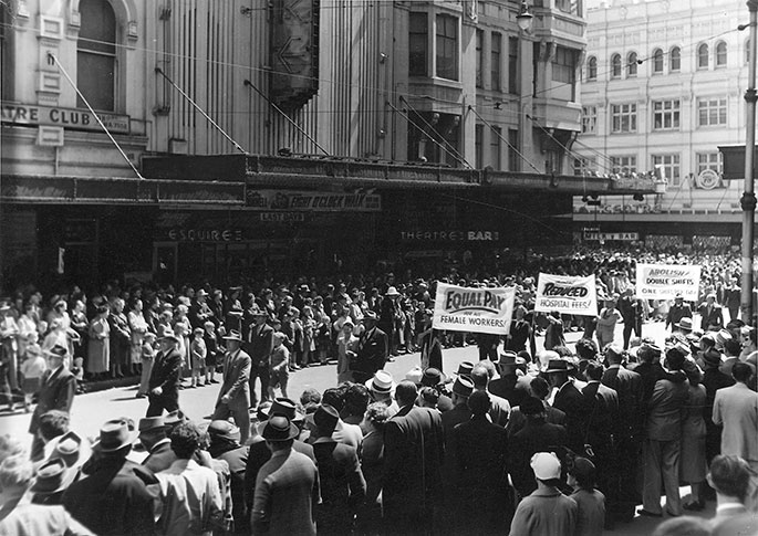 Black and white photo showing men and women marching down street lined with crowds. Workers are holding banners such as Equal pay for female workers.