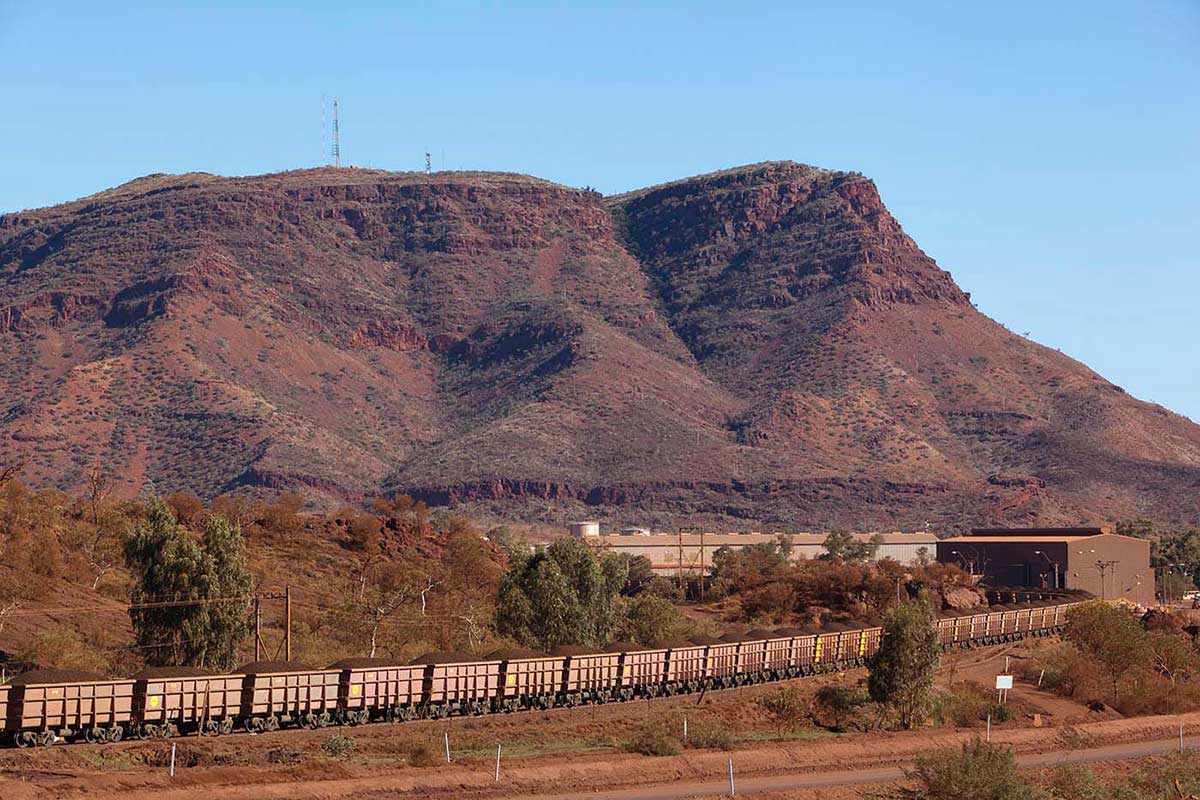Long train with bogeys bulging with iron ore. In background is a low mountain.