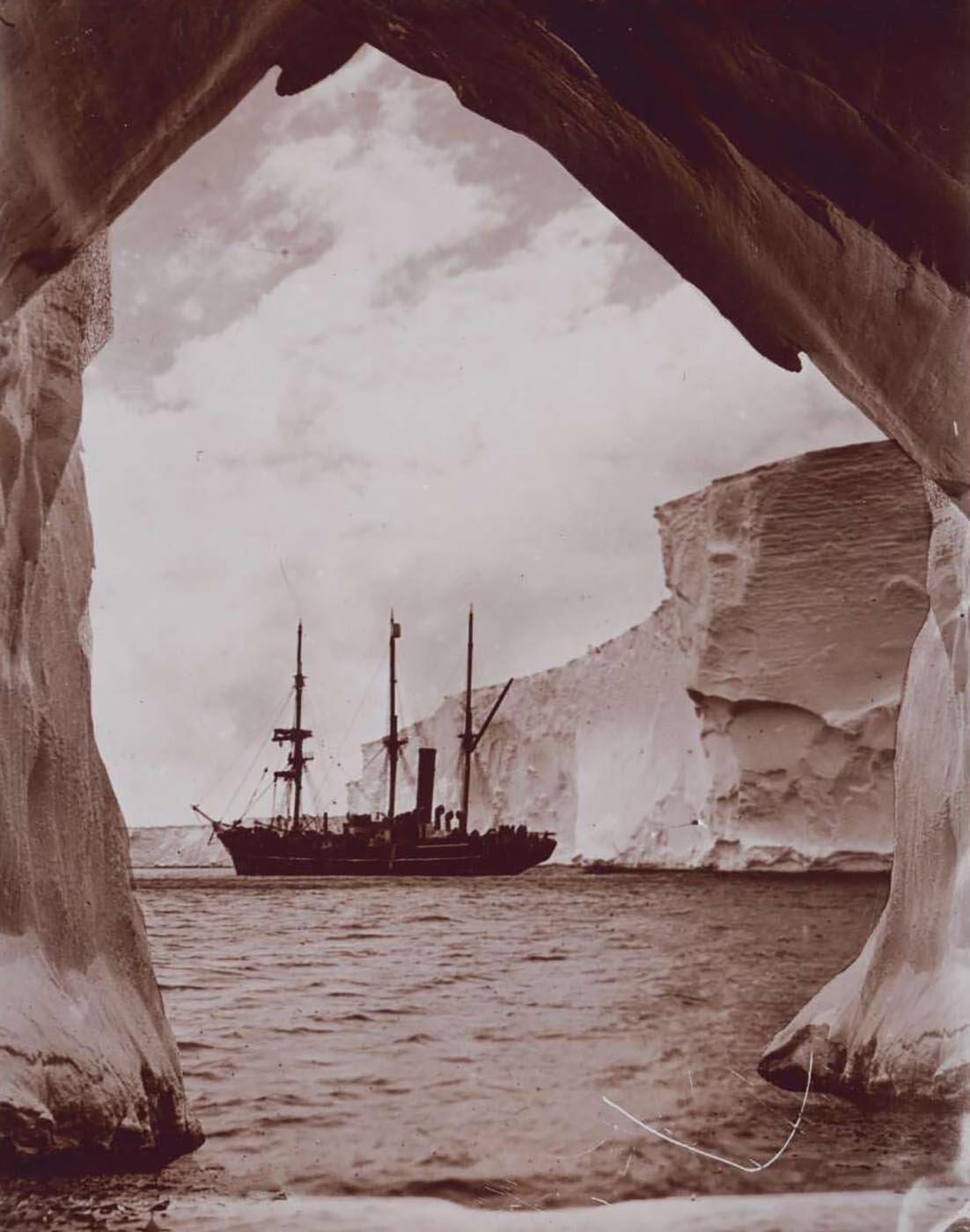 Black and white photograph of a ship spotted through opening in glacial ice. - click to view larger image